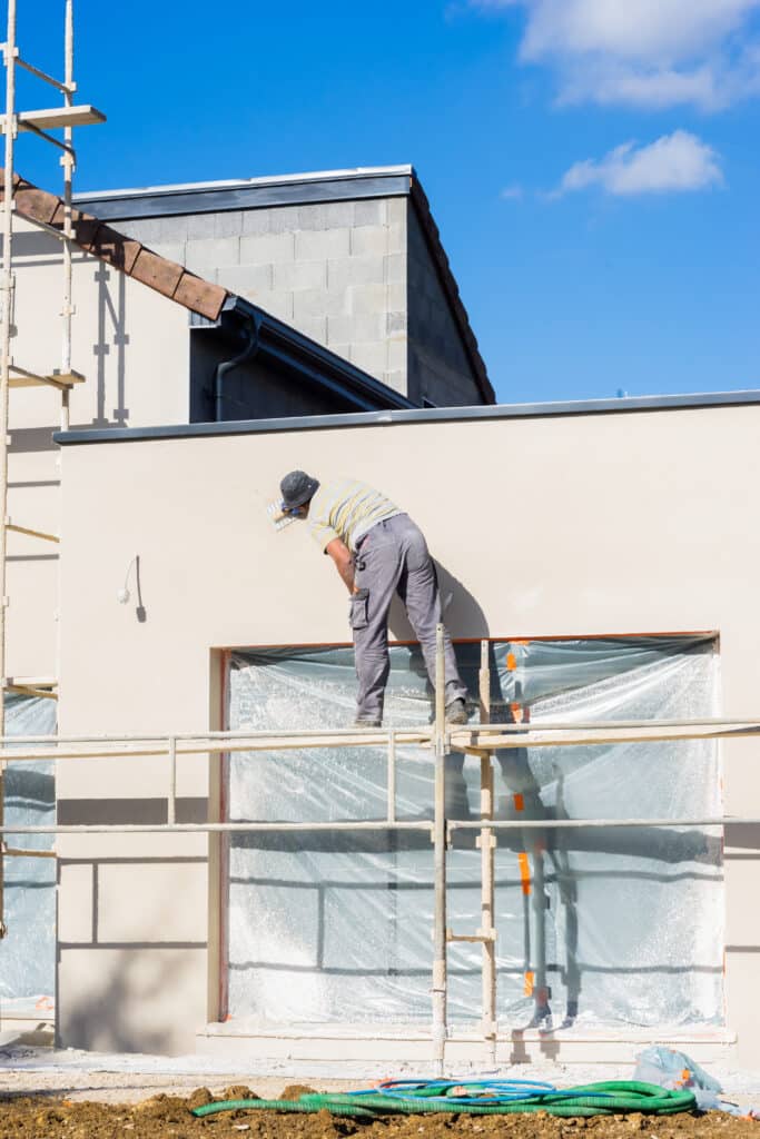 Enduit de façade : l'ouvrier termine la maison neuve. Ouvrier sur échafaudage appliquant l'enduit beige sur la façade d'une maison en construction, sous un ciel bleu intense.
