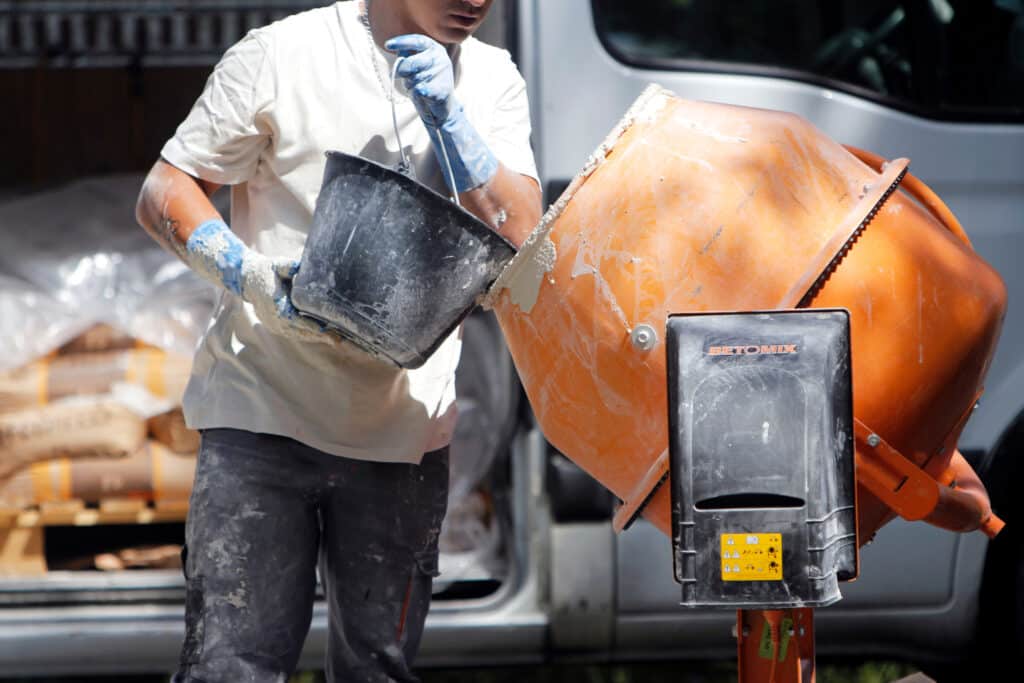 Préparation du ciment : ouvrier charge la bétonnière. Ouvrier chargeant une bétonnière orange BETOMIX avec un seau de mélange. Gants de protection bleus, chantier ciment.